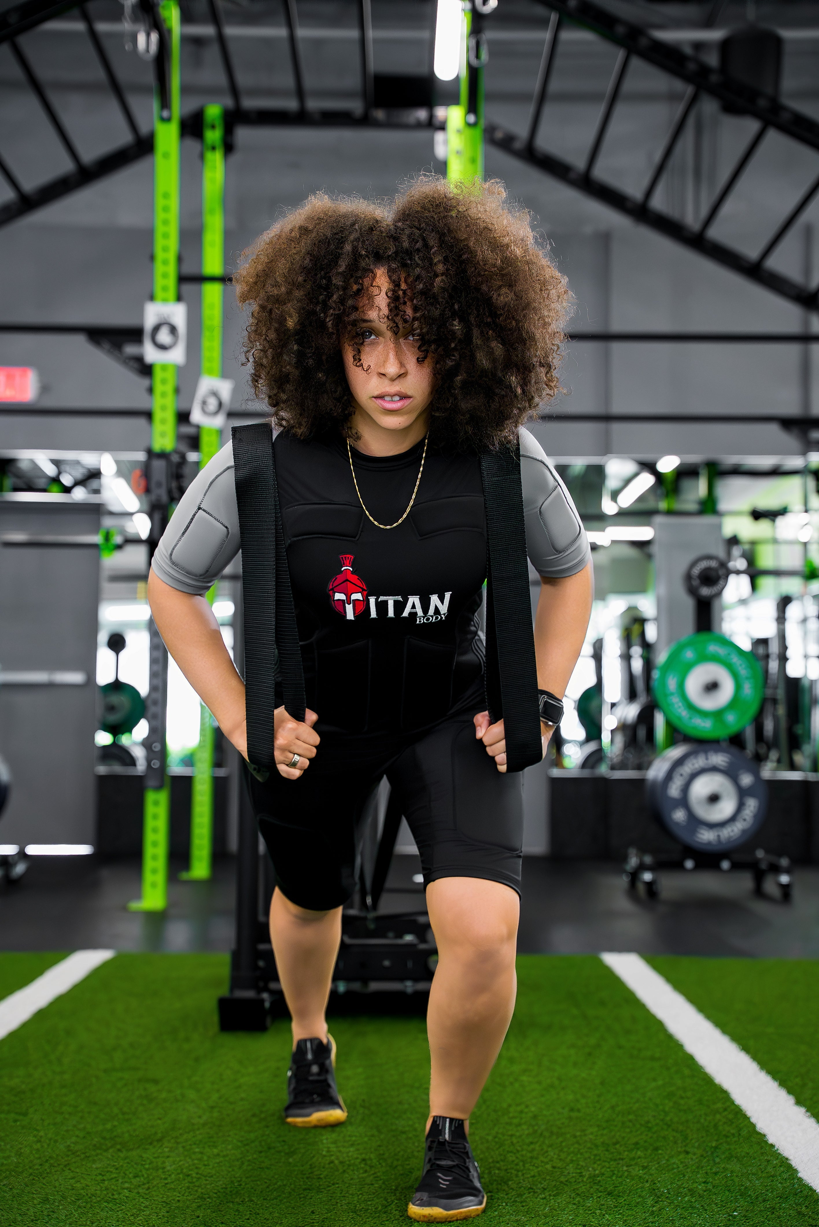 Woman training in Titan Body EMS suits for runners, pulling a weighted sled across turf in a gym