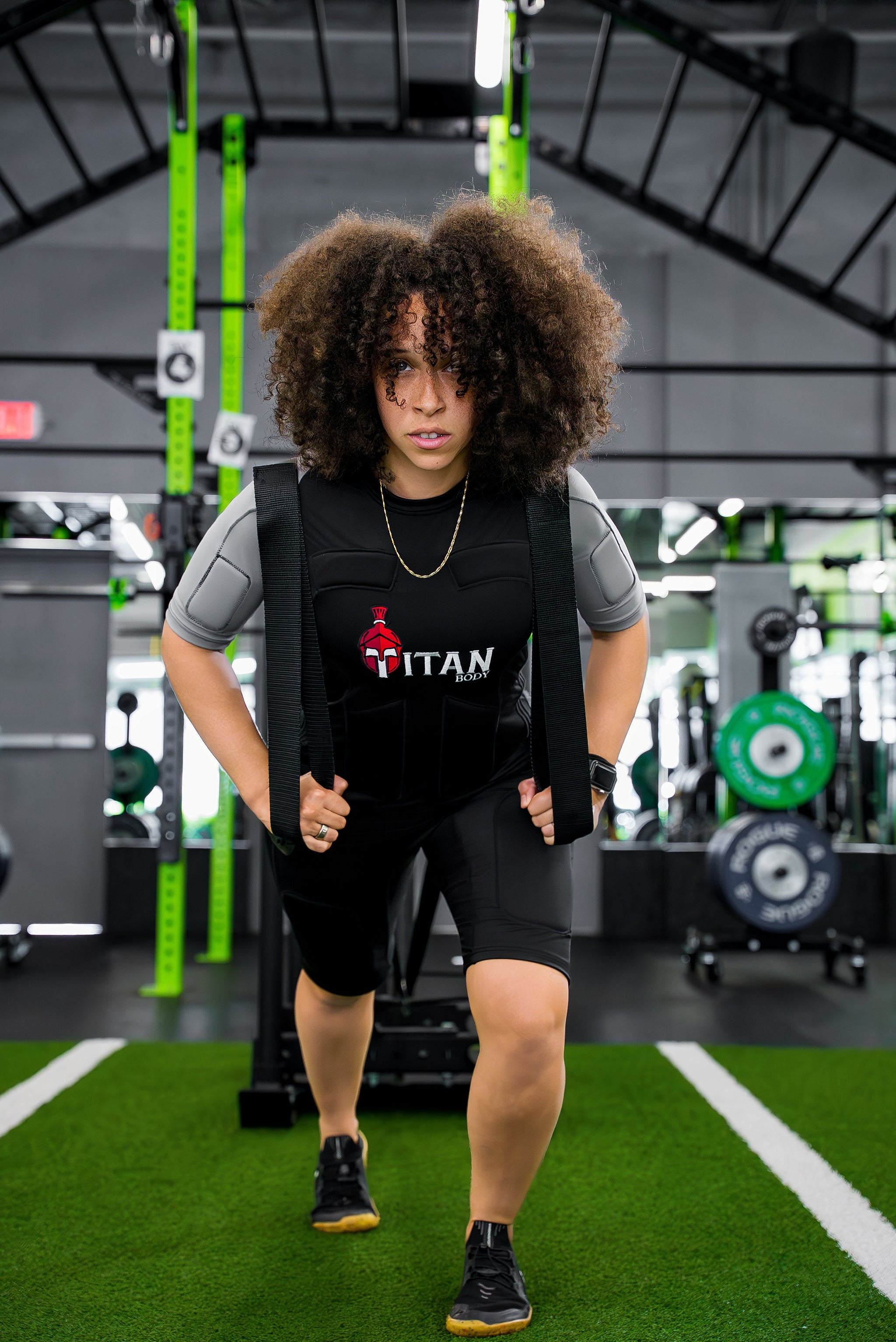 Woman training in Titan Body EMS suits for runners, pulling a weighted sled across turf in a gym