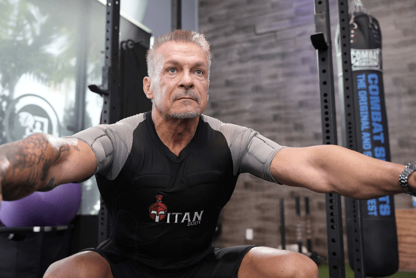 Man wearing a TitanBody EMS suit for weight loss during a squat exercise in a home gym.