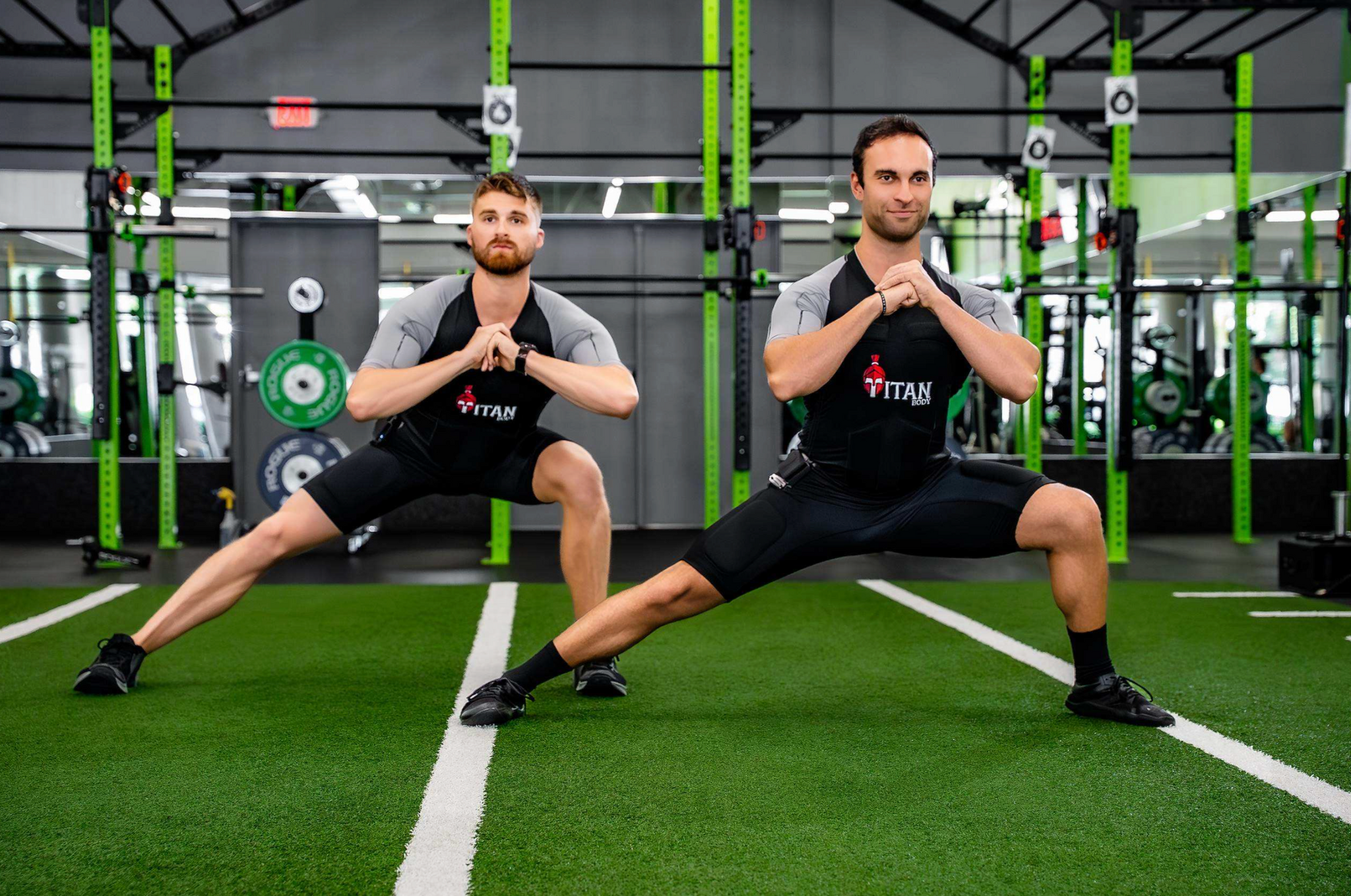 Two men doing side lunges in a gym and in their TitanBody EMS exercise suit.